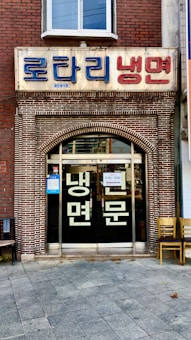 A storefront with a brick facade and a large sign in Korean characters above the entrance. There are glass doors with more Korean writing and a couple of chairs placed outside on the tiled pavement.