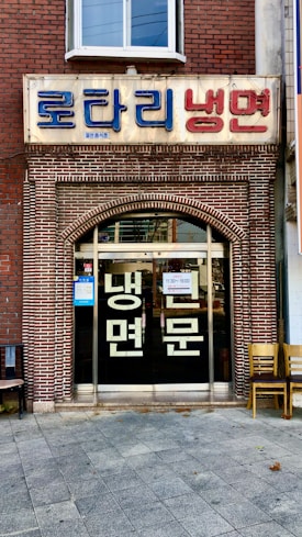 A storefront with a brick facade and a large sign in Korean characters above the entrance. There are glass doors with more Korean writing and a couple of chairs placed outside on the tiled pavement.