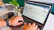 A person is using a laptop displaying a website for designing custom t-shirts. The hands on the keyboard suggest active engagement, with a slice of chocolate cake on a plate and a cup of coffee nearby. A vase with green leaves adds a touch of nature to the scene. A watch is visible on the person's wrist.