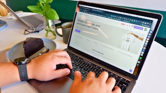 A person is using a laptop displaying a website for designing custom t-shirts. The hands on the keyboard suggest active engagement, with a slice of chocolate cake on a plate and a cup of coffee nearby. A vase with green leaves adds a touch of nature to the scene. A watch is visible on the person's wrist.