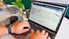 A person is using a laptop displaying a website for designing custom t-shirts. The hands on the keyboard suggest active engagement, with a slice of chocolate cake on a plate and a cup of coffee nearby. A vase with green leaves adds a touch of nature to the scene. A watch is visible on the person's wrist.