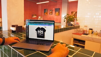 A laptop is placed on a green-tiled countertop displaying an image of a black T-shirt with '404 Not Found' text. The surrounding area includes a plant, some decorative art on pink walls, and kitchen items such as napkins and a coffee machine.