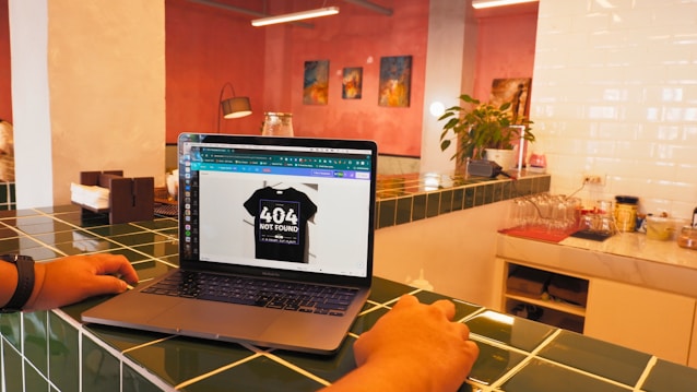 A laptop is placed on a green-tiled countertop displaying an image of a black T-shirt with '404 Not Found' text. The surrounding area includes a plant, some decorative art on pink walls, and kitchen items such as napkins and a coffee machine.