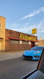 A vibrant blue sports car is parked in front of a brick building with a large sign that reads 'Ridgewood.' The sign is in bold, yellow lettering. There's a person standing near the building's entrance under the clear blue sky.