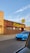 A vibrant blue sports car is parked in front of a brick building with a large sign that reads 'Ridgewood.' The sign is in bold, yellow lettering. There's a person standing near the building's entrance under the clear blue sky.