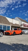 An orange Mercedes-Benz sports car is parked on a paved lot in front of a building with the sign 'Lavallette Clubhouse'. Other luxury cars, including a white Bentley and a silver Ferrari, are also parked nearby. The sky is clear with a few scattered clouds.