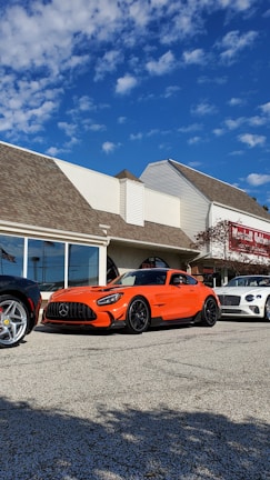 An orange Mercedes-Benz sports car is parked on a paved lot in front of a building with the sign 'Lavallette Clubhouse'. Other luxury cars, including a white Bentley and a silver Ferrari, are also parked nearby. The sky is clear with a few scattered clouds.