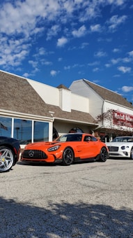 An orange Mercedes-Benz sports car is parked on a paved lot in front of a building with the sign 'Lavallette Clubhouse'. Other luxury cars, including a white Bentley and a silver Ferrari, are also parked nearby. The sky is clear with a few scattered clouds.