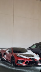 A red sports car with a sleek design partially covered in snow. It is parked next to a black SUV in an indoor setting with beige walls.