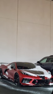 A red sports car with a sleek design partially covered in snow. It is parked next to a black SUV in an indoor setting with beige walls.