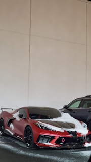 A red sports car with a sleek design partially covered in snow. It is parked next to a black SUV in an indoor setting with beige walls.