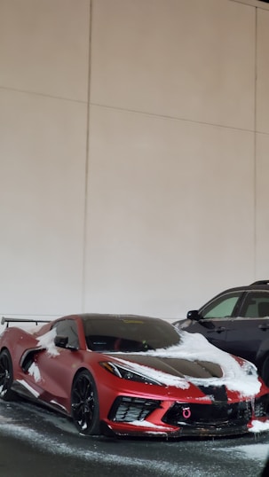 A red sports car with a sleek design partially covered in snow. It is parked next to a black SUV in an indoor setting with beige walls.