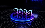 A bold and futuristic representation of the year 2024 is set against a dark, metallic background with circular patterns. The numbers are illuminated with a gradient of blue and purple lights, creating a reflective effect on the glossy surface below.