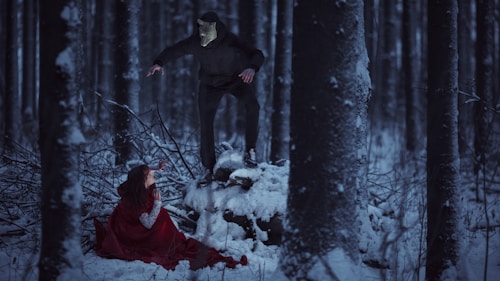 A mysterious forest scene with snow-covered ground and trees shrouded in darkness. A person in a red cloak is crouched on the ground, looking up at a figure in black with a mask standing on a snow-covered log. The forest creates a stark and tense atmosphere.