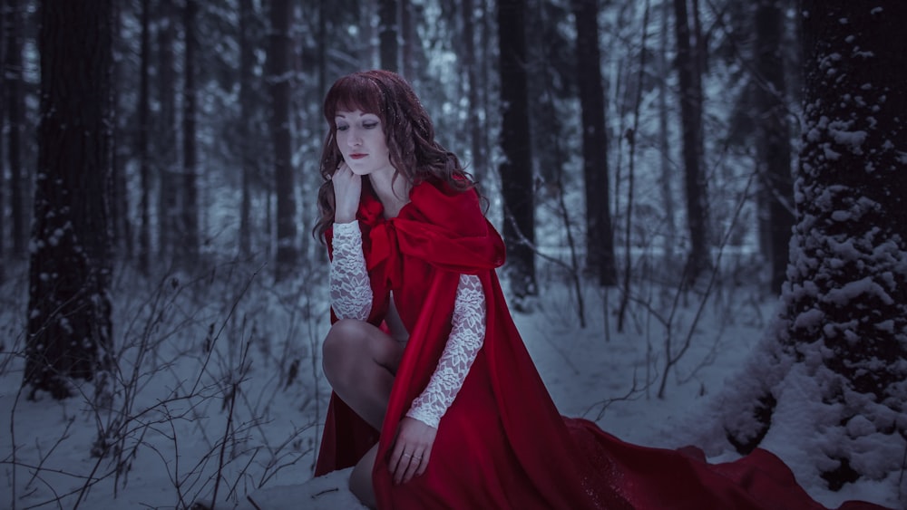 a woman in a red dress crouches in a snowy forest