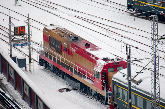 A diesel locomotive is parked on snowy railway tracks. It is painted in shades of red and brown, with visible snow accumulation. Overhead electric lines and a digital sign showing the number 14 are present. A passenger train with dark green cars is adjacent.