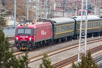 A red locomotive pulling a series of passenger train cars travels along a railway track. The train is surrounded by urban and industrial infrastructure, with overhead electrical lines and utility poles visible. The background includes buildings and bare trees, suggesting a setting in a rail yard or a transit area.