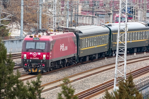 A red locomotive pulling a series of passenger train cars travels along a railway track. The train is surrounded by urban and industrial infrastructure, with overhead electrical lines and utility poles visible. The background includes buildings and bare trees, suggesting a setting in a rail yard or a transit area.
