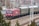 A red locomotive pulling a series of passenger train cars travels along a railway track. The train is surrounded by urban and industrial infrastructure, with overhead electrical lines and utility poles visible. The background includes buildings and bare trees, suggesting a setting in a rail yard or a transit area.