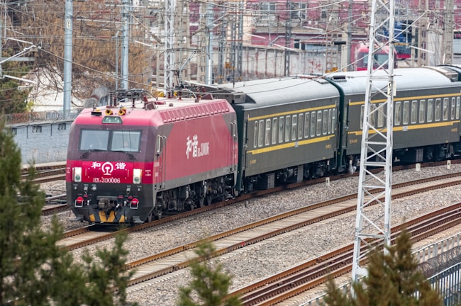 A red locomotive pulling a series of passenger train cars travels along a railway track. The train is surrounded by urban and industrial infrastructure, with overhead electrical lines and utility poles visible. The background includes buildings and bare trees, suggesting a setting in a rail yard or a transit area.