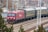 A red locomotive pulling a series of passenger train cars travels along a railway track. The train is surrounded by urban and industrial infrastructure, with overhead electrical lines and utility poles visible. The background includes buildings and bare trees, suggesting a setting in a rail yard or a transit area.