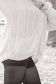 A person wearing a textured, knitted cardigan with cable patterns and buttons, paired with dark leggings. The focus is on the clothing details, including the pocket and the knit stitching.