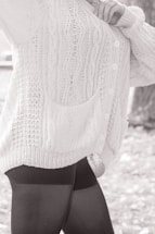 A person wearing a textured, knitted cardigan with cable patterns and buttons, paired with dark leggings. The focus is on the clothing details, including the pocket and the knit stitching.