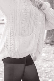 A person wearing a textured, knitted cardigan with cable patterns and buttons, paired with dark leggings. The focus is on the clothing details, including the pocket and the knit stitching.