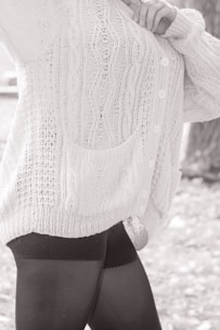 A person wearing a textured, knitted cardigan with cable patterns and buttons, paired with dark leggings. The focus is on the clothing details, including the pocket and the knit stitching.