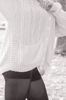 A person wearing a textured, knitted cardigan with cable patterns and buttons, paired with dark leggings. The focus is on the clothing details, including the pocket and the knit stitching.