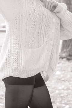 A person wearing a textured, knitted cardigan with cable patterns and buttons, paired with dark leggings. The focus is on the clothing details, including the pocket and the knit stitching.