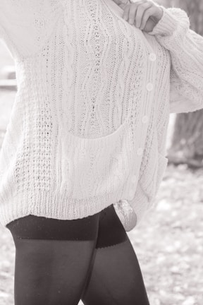 A person wearing a textured, knitted cardigan with cable patterns and buttons, paired with dark leggings. The focus is on the clothing details, including the pocket and the knit stitching.