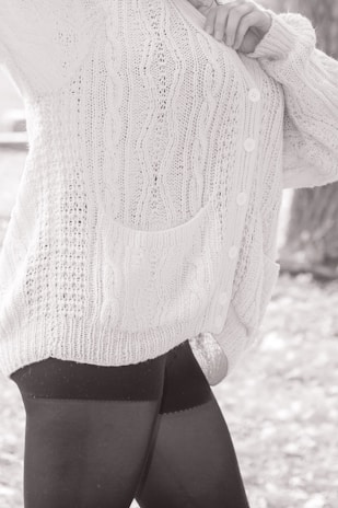 A person wearing a textured, knitted cardigan with cable patterns and buttons, paired with dark leggings. The focus is on the clothing details, including the pocket and the knit stitching.