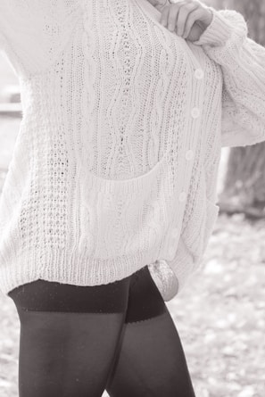 A person wearing a textured, knitted cardigan with cable patterns and buttons, paired with dark leggings. The focus is on the clothing details, including the pocket and the knit stitching.