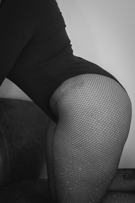 A close-up black and white photograph featuring an individual wearing fishnet stockings and dark clothing. The image focuses on the lower part of the body, captured in a seated position on a soft, cushioned surface.