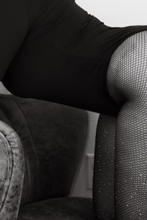 A close-up shot of a person wearing black fishnet stockings and a dark garment, sitting on a plush gray upholstered chair. The image focuses on the textures and patterns of the fabric and stockings.