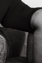 A close-up shot of a person wearing black fishnet stockings and a dark garment, sitting on a plush gray upholstered chair. The image focuses on the textures and patterns of the fabric and stockings.