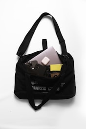 A black shoulder bag with a text design on the front. Inside, there are various electronic items including a laptop, a smartphone with a three-camera setup, a pair of wireless earbuds, and a yellow book with a patterned cover. The bag is placed on a white surface.