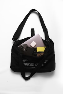 A black shoulder bag with a text design on the front. Inside, there are various electronic items including a laptop, a smartphone with a three-camera setup, a pair of wireless earbuds, and a yellow book with a patterned cover. The bag is placed on a white surface.