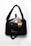 A black shoulder bag with a text design on the front. Inside, there are various electronic items including a laptop, a smartphone with a three-camera setup, a pair of wireless earbuds, and a yellow book with a patterned cover. The bag is placed on a white surface.