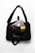 A black shoulder bag with a text design on the front. Inside, there are various electronic items including a laptop, a smartphone with a three-camera setup, a pair of wireless earbuds, and a yellow book with a patterned cover. The bag is placed on a white surface.