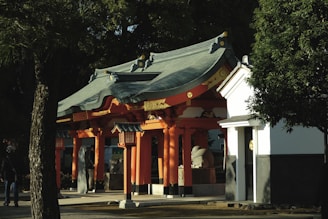 A traditional Japanese shrine features vibrant orange-red wooden structures with a distinct curved roof. Surrounded by lush green trees, the shrine stands out under the soft sunlight, casting gentle shadows across the ground. Lanterns and stone figures add cultural detail to the sacred site.