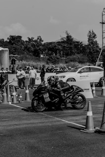 A motorcyclist wearing full gear, including a helmet and protective suit, is positioned in a racing stance on a sport bike. The background shows a white car with 'AMG' branding and several spectators lined up behind a barricade. Traffic cones are placed along the road, indicating a racetrack or event setup. Trees and a clear sky can be seen in the distance.
