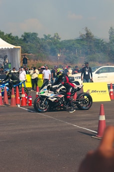 A motorcycle racer in full gear is positioned on a sports bike, ready for a race. The scene is set on a tarmac with traffic cones marking the area. A white car is visible in the background along with spectators, some of whom are taking photos or watching intently. The environment is outdoor, with lush green trees lining the background under a clear sky.