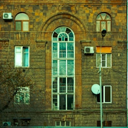 A brick building facade with a large arched window in the center surrounded by smaller rectangular windows. Two air conditioning units are visible, mounted on the wall, as well as a satellite dish. The building's surface has an aged, rustic look with various brown and orange tones. Electrical wires stretch across the scene, and a streetlight pole is positioned in front. Foliage from a nearby tree partially obstructs the bottom corner.