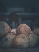 A pile of raw potatoes with rough, dirt-speckled skin lies in a dimly lit environment. The focus is on the potatoes, with a blurred background that suggests indoor storage or a kitchen setting. The lighting casts soft shadows, creating a moody and rustic ambiance.