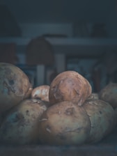 A pile of raw potatoes with rough, dirt-speckled skin lies in a dimly lit environment. The focus is on the potatoes, with a blurred background that suggests indoor storage or a kitchen setting. The lighting casts soft shadows, creating a moody and rustic ambiance.