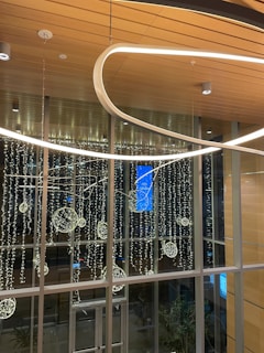 A modern indoor space featuring a high ceiling with wooden panels. Elegant, looped light fixtures hang from above, and illuminated decorative spheres are arranged artistically against a backdrop of large windows. Outside, city lights can be faintly seen through the glass.