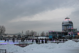 A winter scene with people ice skating on a rink surrounded by snow. There is a large building with a domed roof and observation deck in the background. The sky is overcast, and there are various colorful lights illuminating the skating area. Trees and another architectural structure, possibly a bridge, are visible in the background.