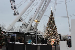 A large decorated Christmas tree adorned with lights and ornaments is set in an outdoor area near a ferris wheel. Snow covers parts of the scene, creating a wintery atmosphere. There are people gathered around, wrapped in winter clothing, suggesting a festive event. Nearby structures and lamp posts are also visible.